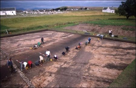 A photograph of a large open area excavation taken from an elevated position showing 14 volunteers in a line with trowels