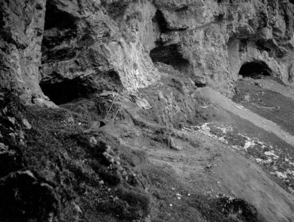 A black and white photograph showing three caves at the foot of vertical cliffs