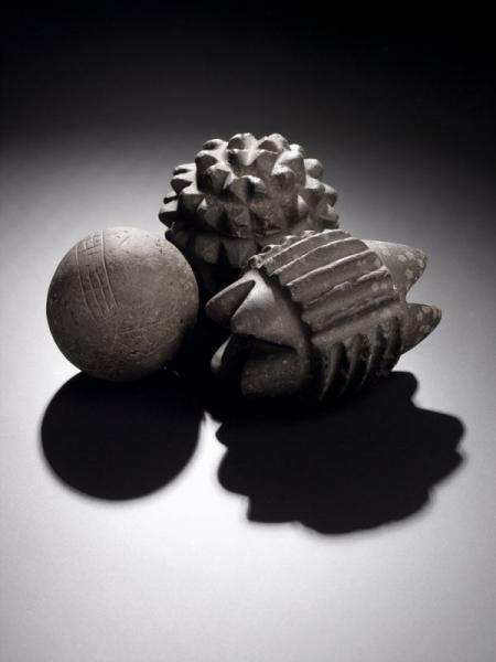 A photograph showing a smooth stone ball with a faint linear decoration, a carved stone ball with protruding pyramids and a cylindrical carved stone with groves along the body and four points at each end