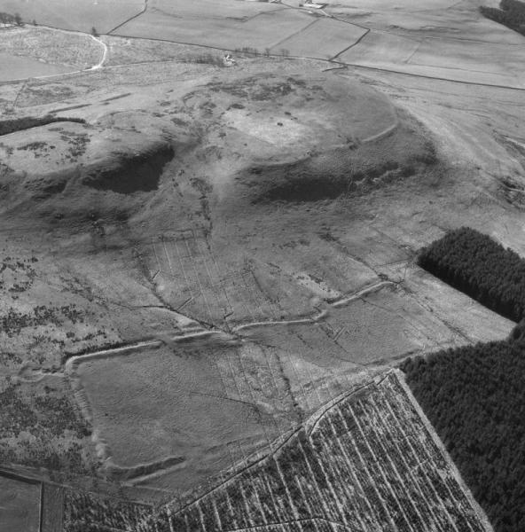 A black and white aerial photograph showing an agricultural landscape with extensive remains of rectilinear earthworks near the base of an imposing hill 