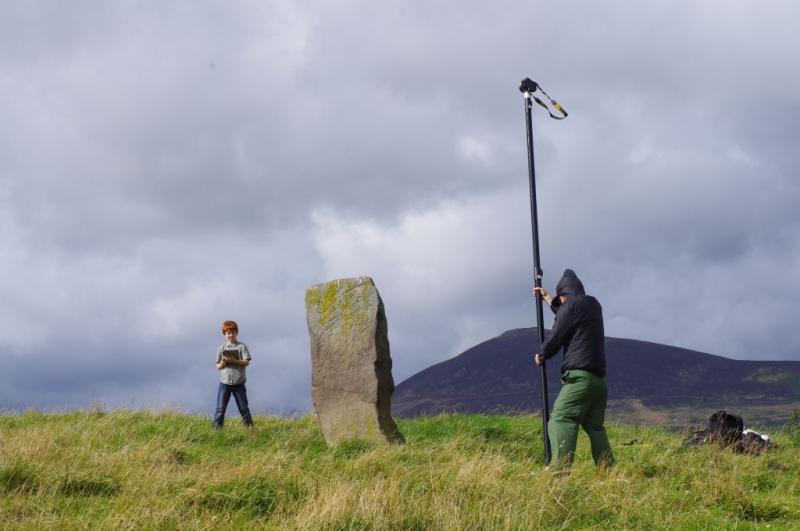 Two people on a grassy hill, one holding a camera on a high pole attempting to take images of a standing stone from many angles