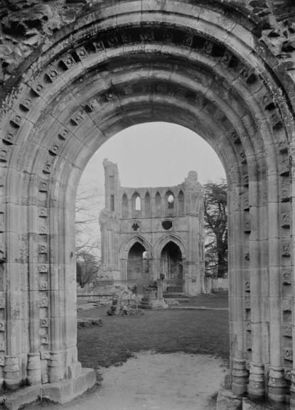 A balck and white photo of a medieval stone arch with the remains of a ruined abbey in seen through the doorway