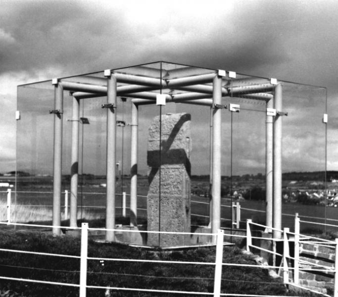 A black and white photo of a standing stone slab stood in the country side. The stone is surrounded by a steel frame supporting a glass cube