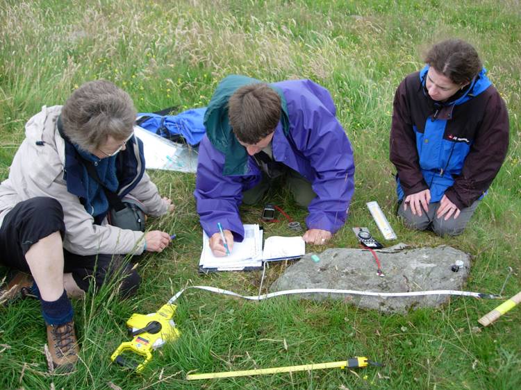 Three people sat on the grass around a small carved stone, using measuring tape and a compass to record details on a clipboard