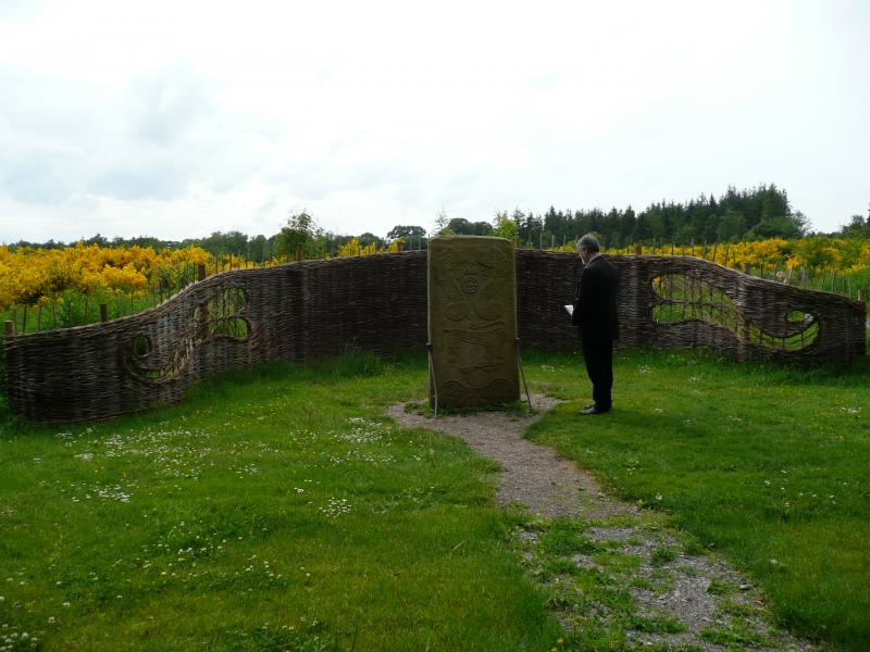 A colour photo of a man stood in front of a cross slab in a field. Behind it is a curving wall made of willow.