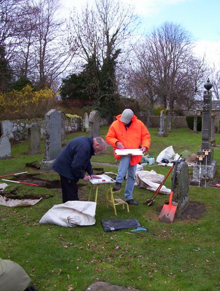A colour photo of two men in a graveyard. They have removed turf from an area with spades and are now recording what they have found on clipboards
