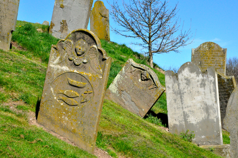 A photograph of old gravestones leaning at different angles on a grassy hillside