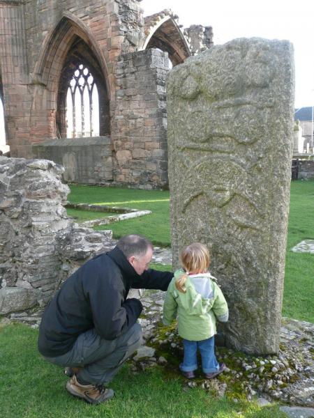 A photograph of a man crouching on the ground pointing out to a little girl details on a large cross slab within a graveyard