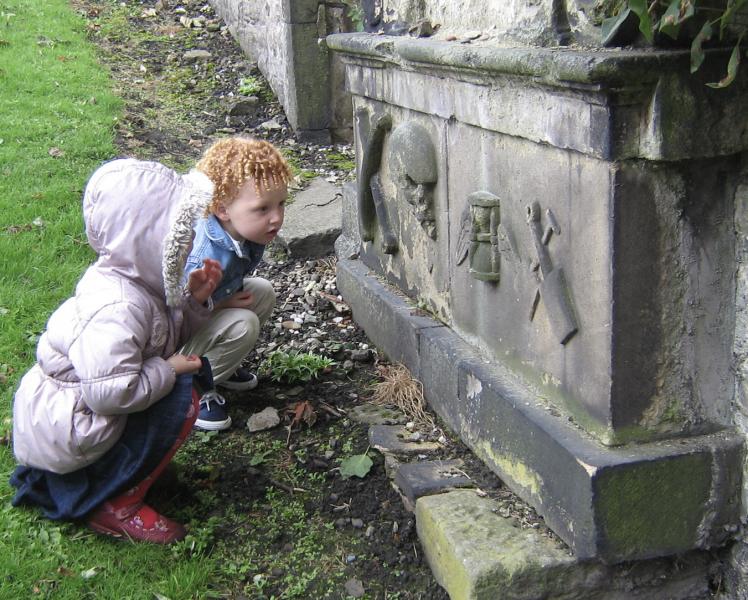 A colour photo of two children kneeling down to get a better look at carvings on a gravestone plinth