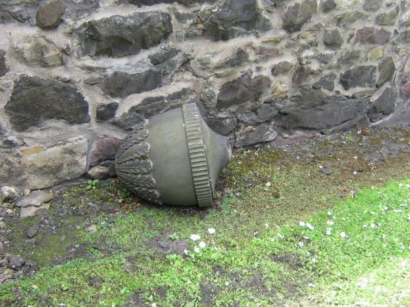 A photo of a large broken top of an acorn shaped stone finial lying on the ground next to a stone wall