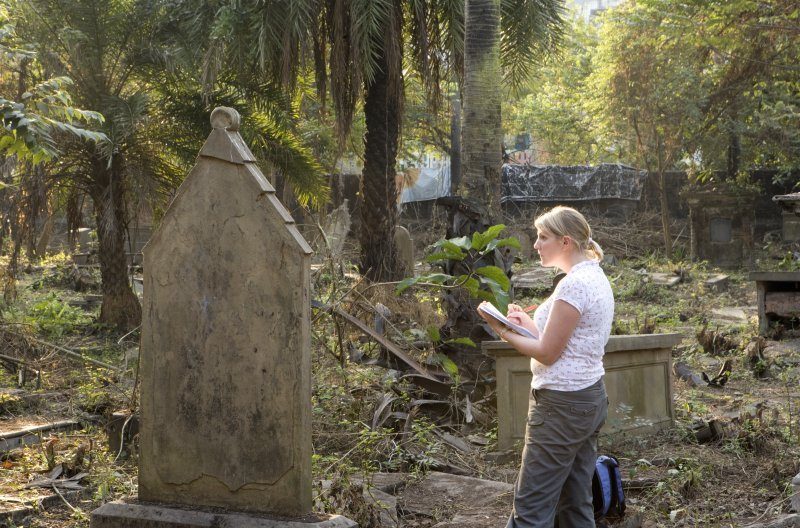 A woman with a clipboard, paper and pencil is looking at a gravestone and taking notes with palm trees in the background