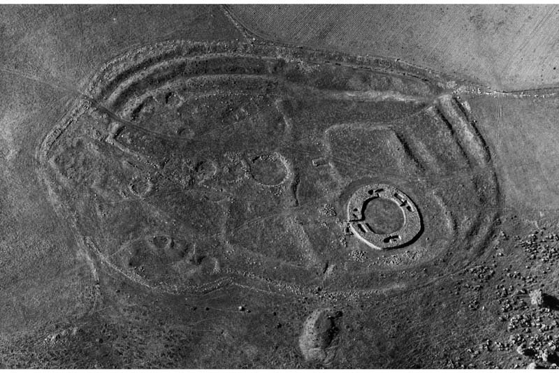 A black and white oblique aerial photograph showing the substantial remains of a circular stone built broch tower with cells and the remains of several roundhouses enclosed within extensive earthworks comprising banks and ditches set within a wider agricultural landscape