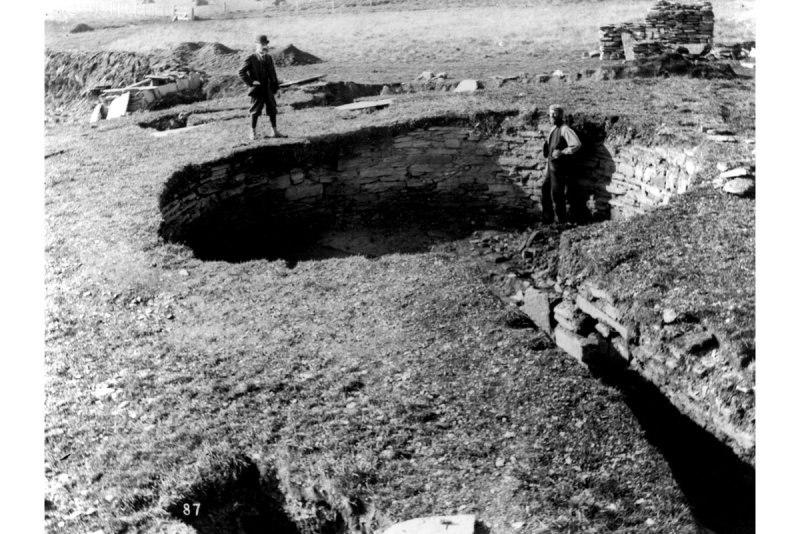 A black and white photograph showing the excavated circular interior of a broch structure with one man standing inside and another on the edge looking down 