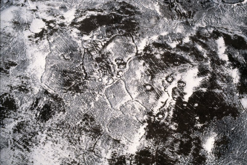 An aerial photograph of a snow covered landscape showing the remains of irregular shaped prehistoric field systems and several stone-walled roundhouses 