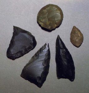 A photograph showing three black flint arrowheads with retouched edges and a smaller brown flint arrowhead and a circular scraper