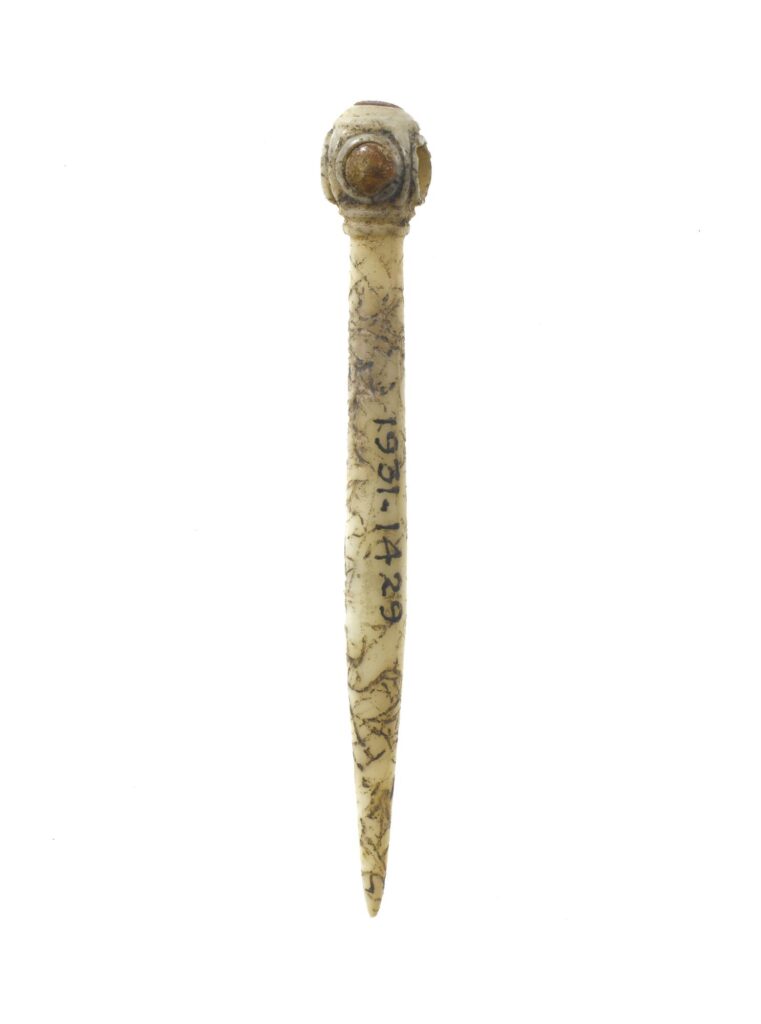 A close up photograph of a yellow-brown bone pine, taken against a white background. The pin has a round head, with a circular piece of orange amber inserted into the head. The shaft of the pin is long, and comes to a point. Brown scratches are visible across the pin, and the item number 1931-1429 is written down the pin shaft in black ink. 