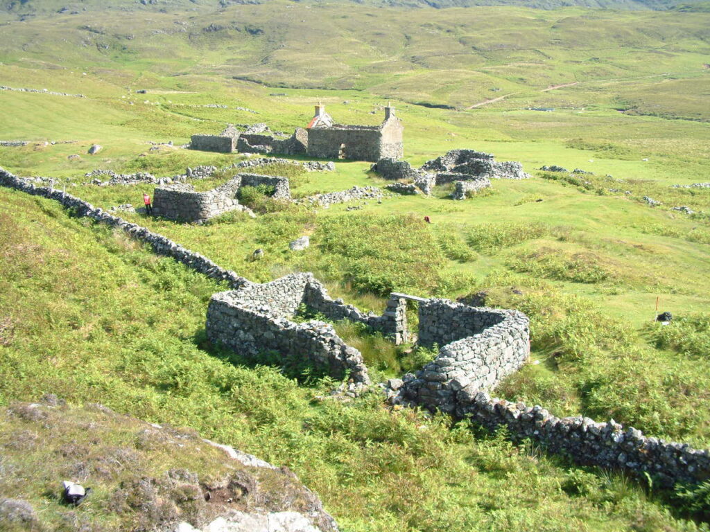 In the foreground sit the stone walls of a rectangular building set into a boundary wall. In the background, the remains of several other stone buildings sit. 