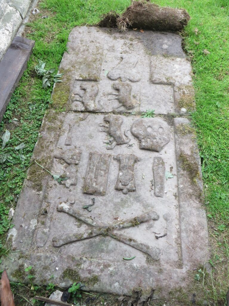 Stone slab on the ground with various symbols on it, including a coffin, skull, crossbones, and a hand holding a bell.