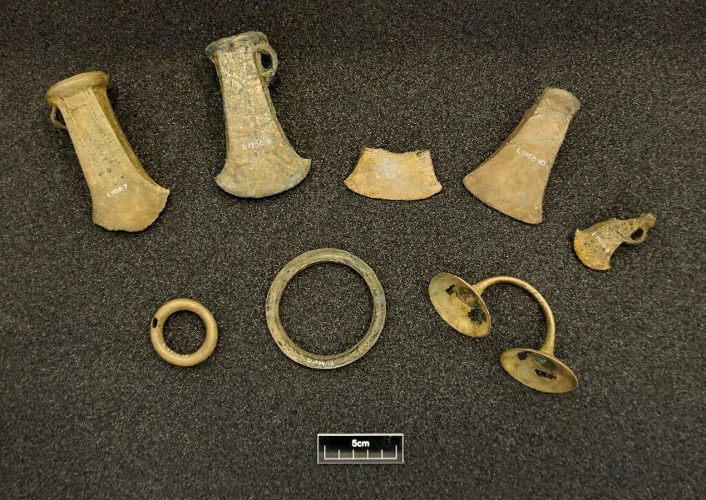 The Poolewe metalwork hoard laid out on a black background. Consists of 8 pieces including 5 axes ranging from fragmented to well-preserved. 5cm ruler for scale