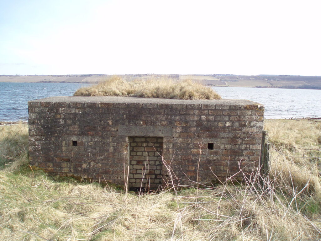 A box-shaped brick building, with the door blocked up, sitting near the shore of the Cromarty Firth.