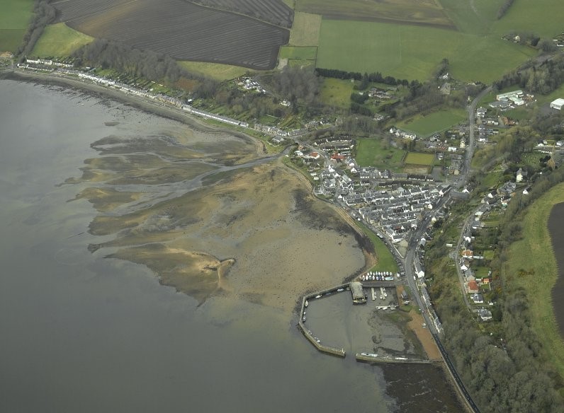 Oblique aerial photograph of a coastal village, with the beach and sea covering half of the image diagonally, to the west and south. To the east and north, a village of light coloured houses and buildings separates the beach from a green landscape. In the foreground a semi-circular harbour, lined with small boats, can be seen, defined by a grey wall curving from the land to the water. The tide looks very low, with sand protruding from most of the visible water. 