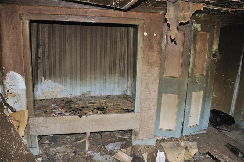 Photograph from inside a derelict houses, facing a box bed, which is made by building a wooden bed platform within an alcove.  The bed and floor are covered in wallpaper, dust and debris. The ceiling has partially collapsed and the rafters are exposed at the top of the image. 