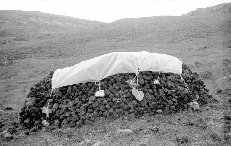 A black and white image of a large pile of lumps of peat, covered in a white tarpollin weighed down by large stones on ropes. 