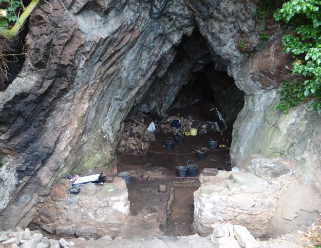 View inside a cave during excavation. The cave is triangular, dark and appears very deep. Buckets, trowels and grid ropes can be seen. 