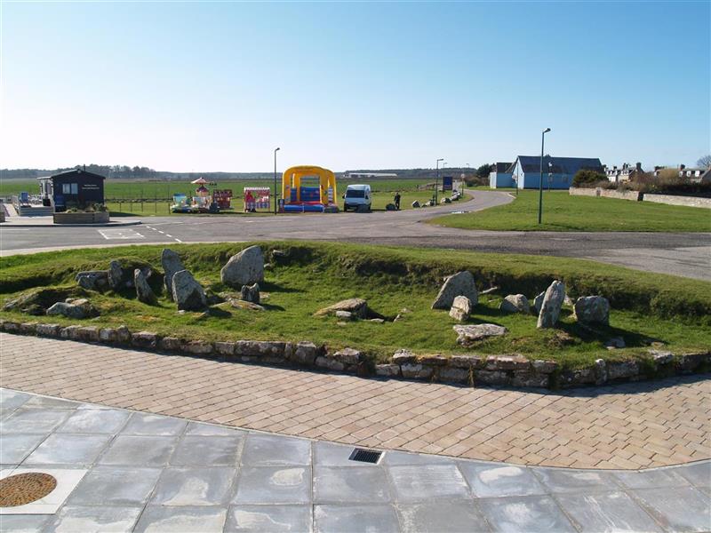 A group of erect stones in a grassy area located next to a car park and a road. 