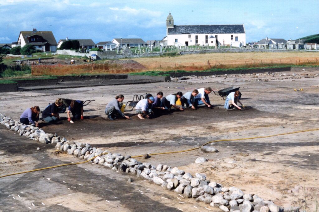 Image shows a horizontal line of ten people, crouched and trowelling in a large, open trench. They are digging in a line, with three wheelbarrows behind them. The soil shows a range of features with light brown and dark brown soil. A line of white buildings can be seen in the background. 