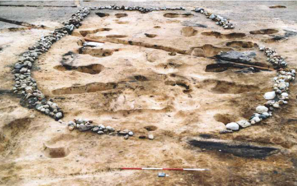 Oblique image showing the excavated remains of a stone-lined structure of sub-oval shape. The line of small stones and pebbles is curved on the side facing the camera, and appears to be squared off on the opposite side. The soil is light yellow and brown, with dark brown areas representing features. Small pits have been dug both inside and outside the structure, appearing to create the shape of postholes which would be used as wall and roof supports for the structure which once stood in this place. 