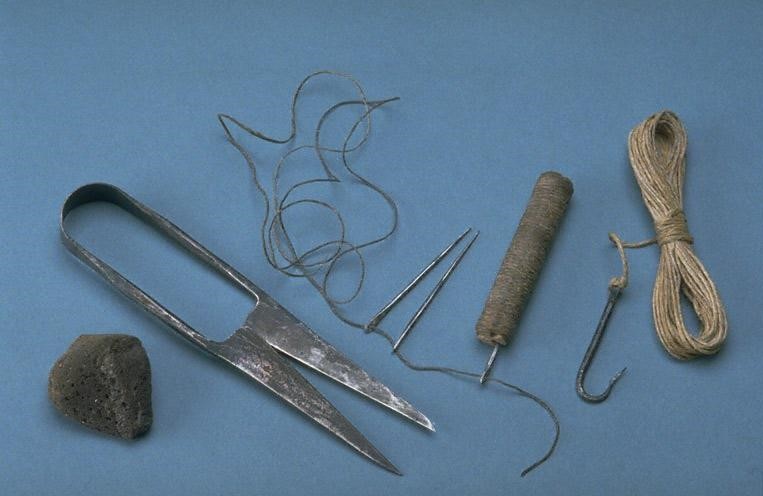 Image of grave goods against a blue background, from above. From left, we see a dark brown pumice stone, irregularly shaped, then a distressed pair of shears which have two triangular blades attached by a thin handle that curves at the top. In the centre in two iron needles, slightly tarnished, with a cylindrical wooden peg with a metal loop attaching it to a long, coiling thread, which is thread through the wooden peg and both needles. To the right of the image is a metal fishing hook of dark grey iron, attached to a neatly wound coil of brown thread. 