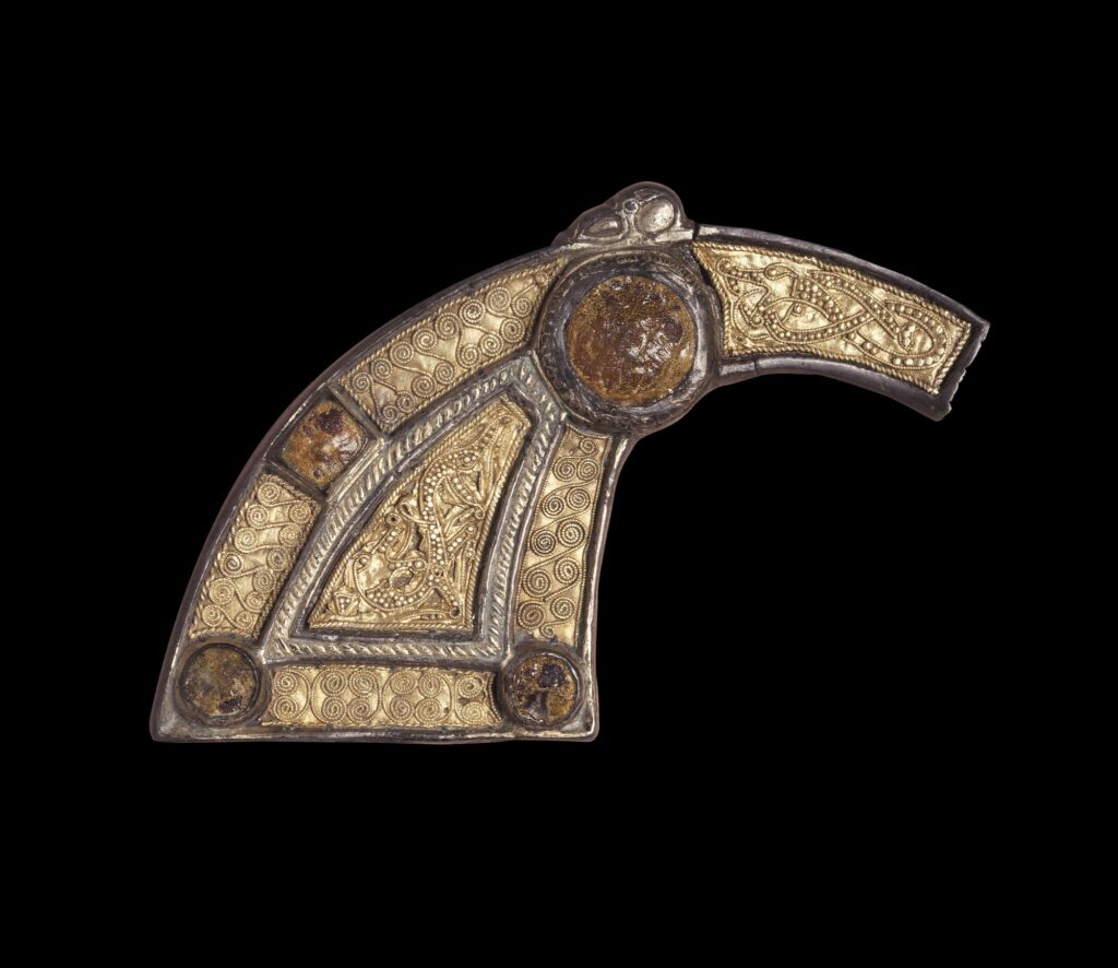 A close-up image of a golden fragment from a pennanular brooch, with orange amber and intricate line designs. Taken against a black background. The artefact is arc shaped, with a thick bottom which thins out towards its top. A border of silver surrounds the gold coloured, patterned area. 