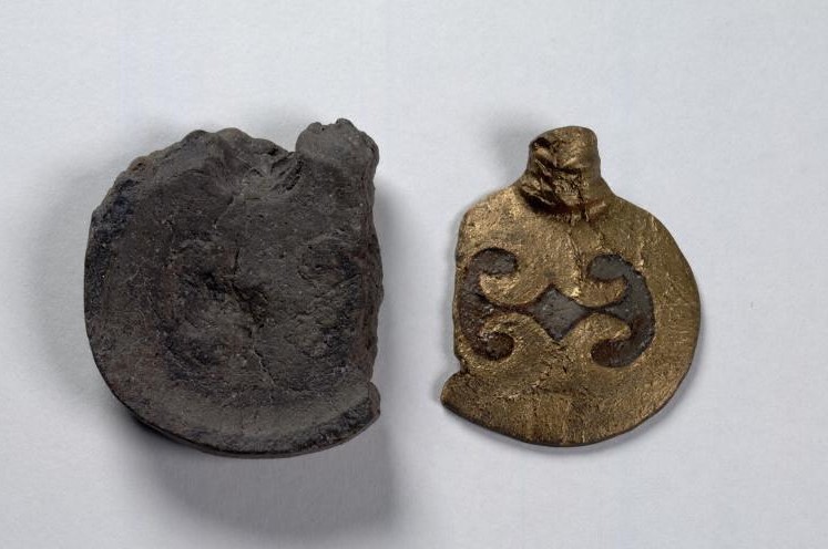 Image of a broken casting mount with two separate pieces, photographed against a white background. One the left is the bottom piece of the mount, which is black and shaped like a bowl, with broken edges on the right and top sides. On the right, a gold coloured piece is the same shape as its counterpart, and a mirror image of the bottom piece. It has a moulded head at the top and a crescent design stamped into it. The piece on the right would be flipped and would fit snuggly into the piece on the left, making a full mount for moulding a bowl. 