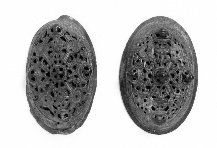 Photograph of oval, intricately designed brooches taken in black and white against a white background. Both brooches are very similar in appearance and size. The brass has been moulded to resemble a lace pattern, with a central sphere and the design creating piers towards the edge of the brooches. Both brooches also have four spheres of brass at the top, bottom and two sides. A plain border surrounds both items. 