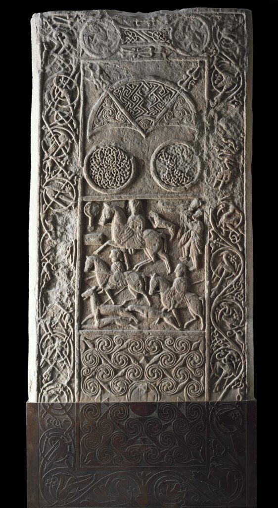 Photograph of a long, rectangular, symbol stone which stands vertically against a black background. The stone is a light brown to grey colour and the carving is done in relief. A border surrounds the edge of the stone, filled with curved decorations and celtic designs. The central area is split into three equal sections. The top section shows a cresent and v-rod symbol, with two circular designs below it, filled with intricate lines. The middle section depicts a battle scene, with men on horses. A small mirror and comb is carved in the top left hand corner of this scene. The bottom section is filled with curving designs, intertwined and swirling. The stone is incomplete at the bottom, with a metal plate holding it in place and completeing the bottom section of the design as it probably appeared originally. 