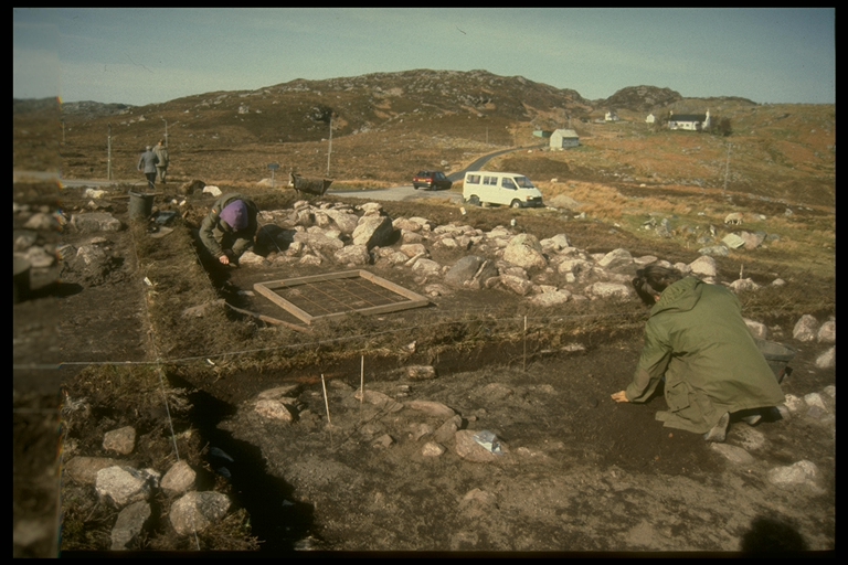 Photograph of an ongoing excavation showing two people trowelling, one in the foreground and one in the midground. The photograph shows a bright, sunny day. The site has large, white boulders throughout and is located at the bottom of hills, seen in the background. 