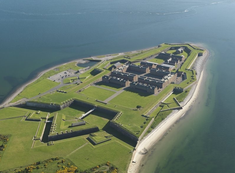 Oblique aerial photograph of a green outlet of land, surrounded by a narrow beach and water. The image shows the elevation of structures from the green, well trimmed grass, with a surrounding wall and angular ramparts. Within the ramparts are rows of rectangular buildings, themselves with a central court area. The walls and structures are all very angular, many forming triangles and angled alcoves within the rampart walls. 