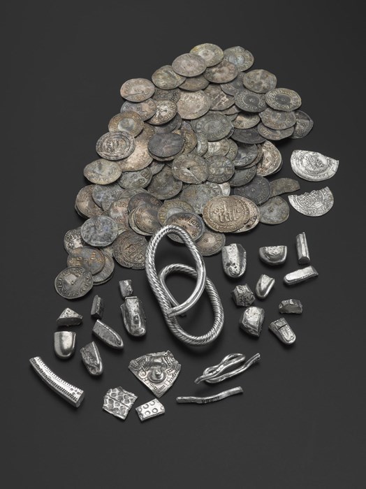 Photograph showing a collection of silver artefacts, with a bundle of coins of varying sizes, all of which are tarnished and have taken on a dark grey to brown colour. In the foreground, highly polished silver items are seen, including small pieces of irregularly shaped silver. In the centre is a silver armlet, or bracelet, made of one, long piece of moulded silver. The items are photographed against a black background. 