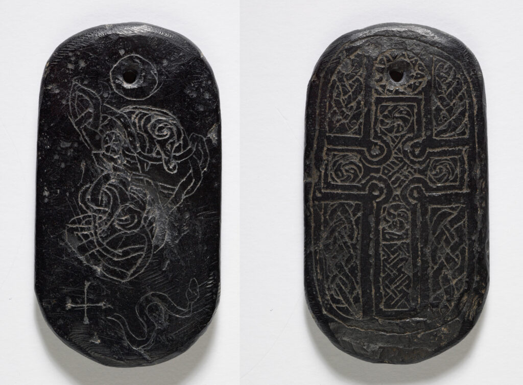 Photograph shows both sides of a carved jet pendant. The pendant is sub-rectangular with rounded ends at the top and bottom. A small hole at the top of the pendant is irregularly shaped. On the left, the front of the pendant is carved with an intricate, interlaced design, as well as a serpent and thin cross. The hole has a thin line carved around it. On the right, the reverse of the pendant shows a Christian cross, folled with an intricate celtic design. The background is also interlaces, and the hole at the top is surrounded by triangular shapes, resembling a sun symbol. 