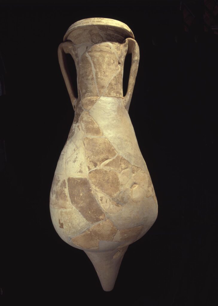 A yellow-brown vase with long, narrow handles at the top, a rounded lower half and a pointed base. It is presented against a black background. Evidence of refitting and repair are clear with cracks and filled lines. 