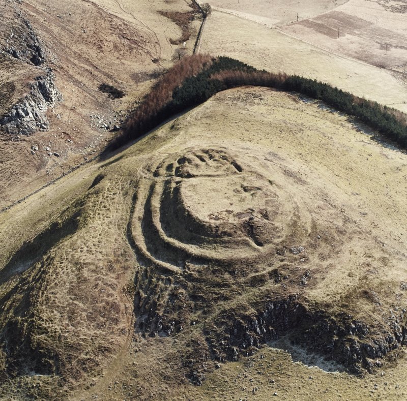 Aerial oblique photograph of an oval hillfort, showing the surrounding landscape of open land and limited trees. The fort has three levels which are separated by step-like gradients, with the top level having bumps and features still visible. 