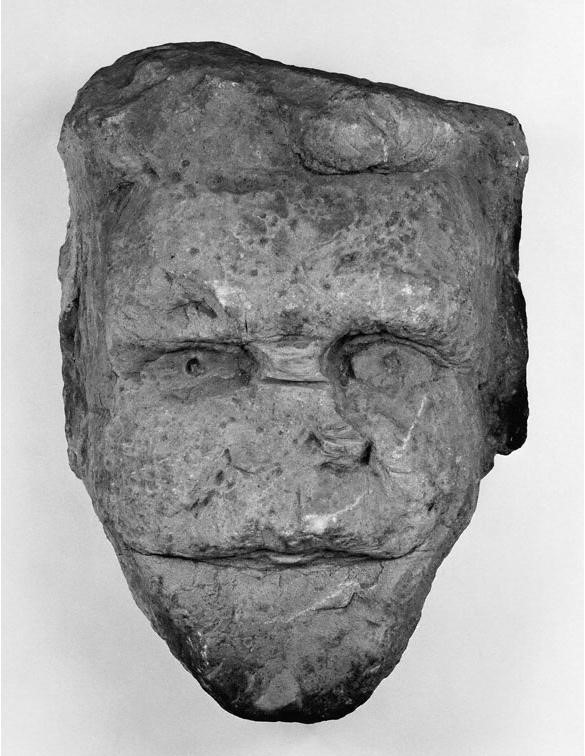 Robust sculpture of a man's face, with deep set eyes, a broken off nose and simplified mouth. His head is square at the top and he has a large chin and jaw area. The sculpture is worn and chipped in many places. 