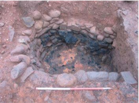 Oblique photograph of a truncated oval subterranean structure, with one straight side. The scooped feature is lined with different sizes of cobbles and the bottom is natural soil. 