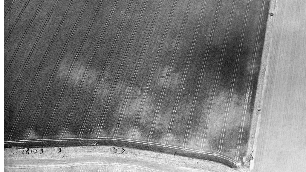 Black and white aerial photograph of a field with a circular cropmark in the centre, and, to its right, dark spots or pits are seen. 