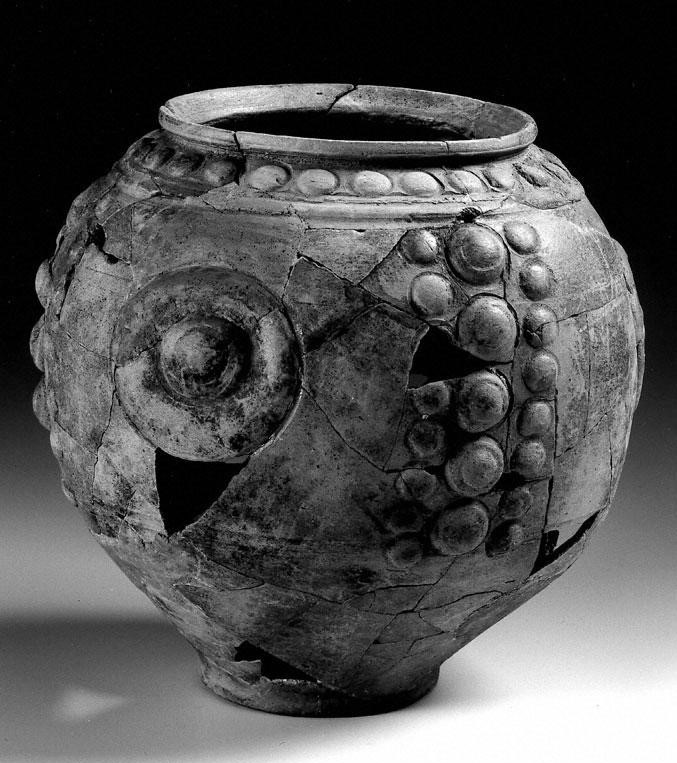 A black and white photo of a rounded vase-type object. Decorations of raised spheres are placed around the lip, as well as concentrated in areas on the body of the vessel. 
