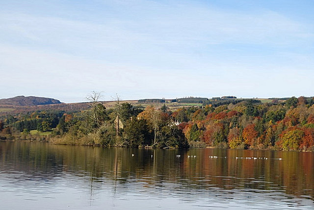 Image of a dense forested landscape taken across a large loch. The trees are green, orange and yellow. A castle or large medieval building can be seen amongst the trees. 