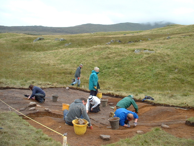 Image of seven people excavating an open trench in a hilly landscape. They have trowels, knee pads and buckets. The sky is grey and overcast. 