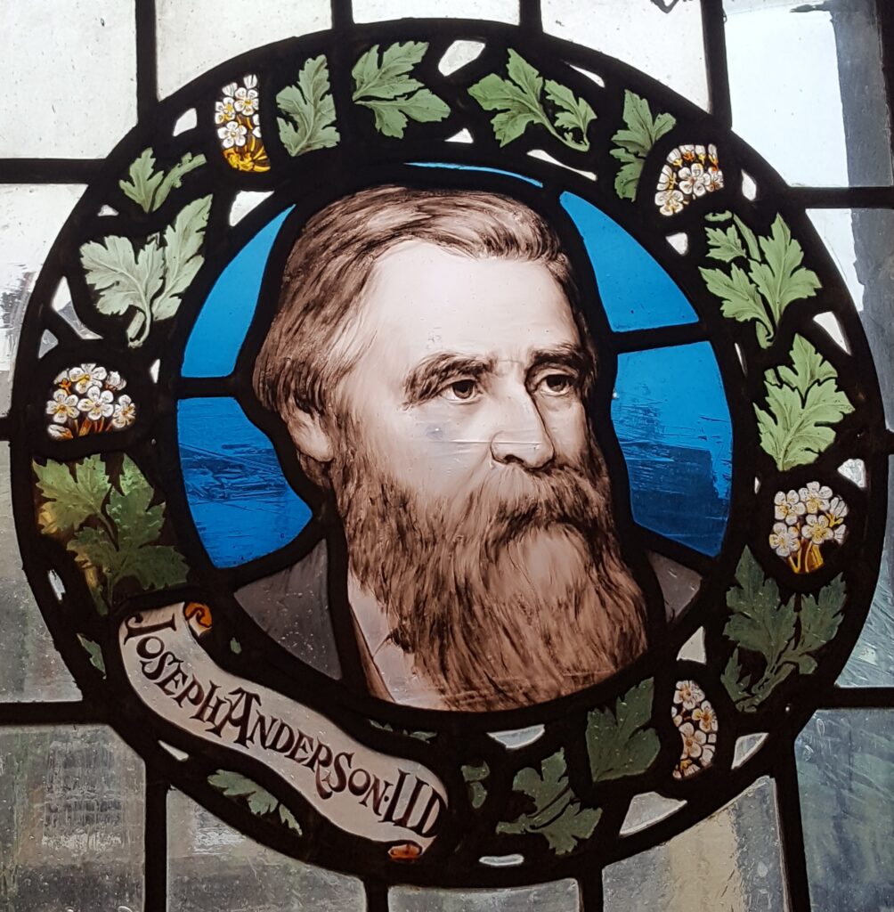 Coloured portrait of a bearded man with brown-red hair looking off into the distance on a stained glass window. His head and shoulders are encircled by a stained glass wreath with leaves and flowers. His name, Joseph Anderson, is written on the wreath. 