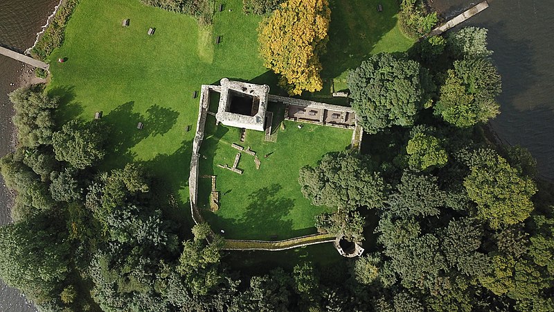 Aerial image of a partially collapsed building in a courtyard built of the same white stone. The main building is square and the courtyard is square with one curved corner. The castle is surrounded by trees. 